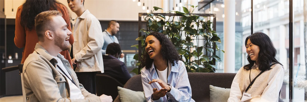 En man sitter i en rullstol och samtalar med två kvinnor som sitter i en soffa. Bakom syns fyra personer. 