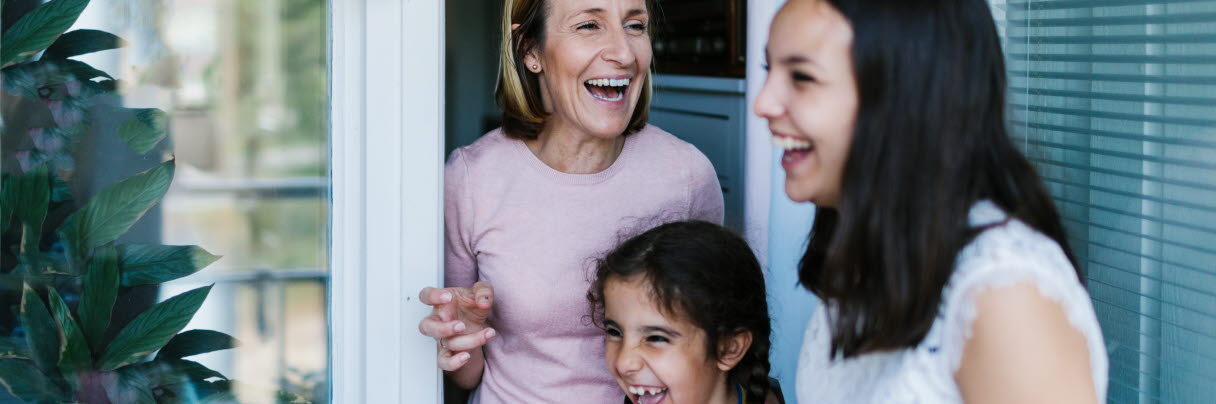 En äldre kvinna och två tjejer står i en balkongöppning och skrattar. 