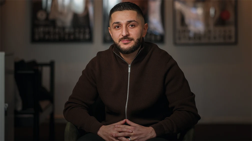 Man med mörkt hår och skägg, i brun tröja som sitter i en fåtölj. 