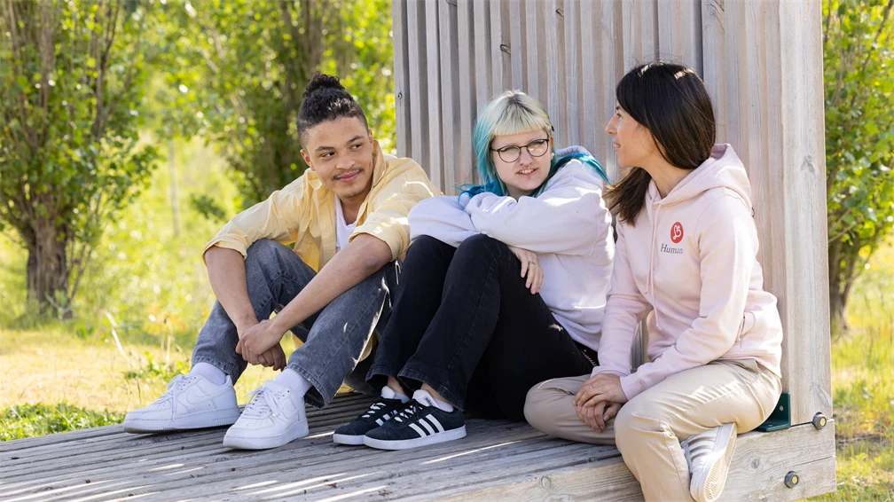 En kvinna och två ungdomar sitter på en träställning, de tittar på varandra