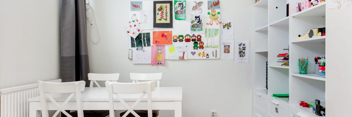 Vitt rum med bord och stolar i vitt. På väggen hänger handmålade konstverk från elever. 