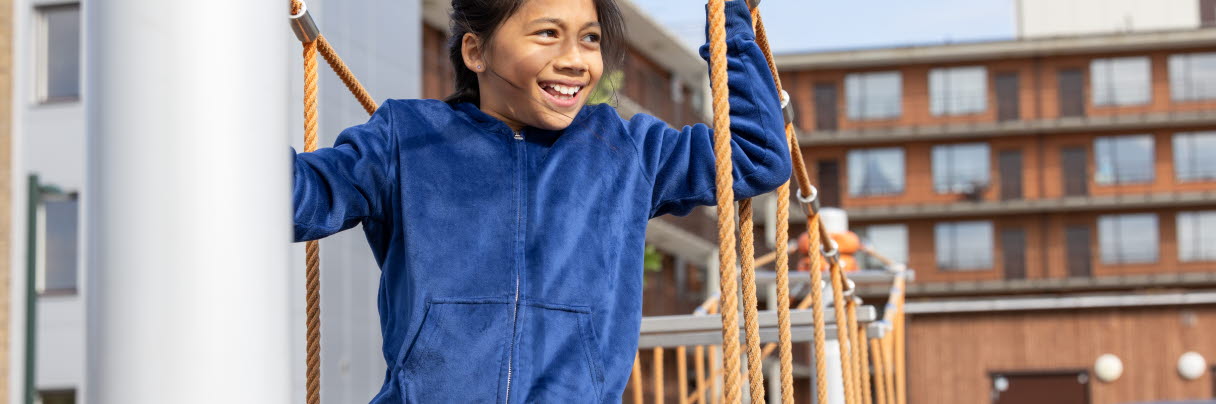 En flicka med mörkt hår och blåtröja står i en klätterställning, hon är glad 