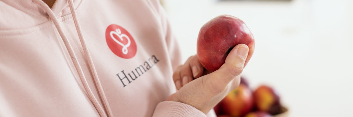 Kvinna iklädd rosa hoodie med Humana logotyp på. I bild syns bröstkorg, i handen håller hon ett rött äpple
