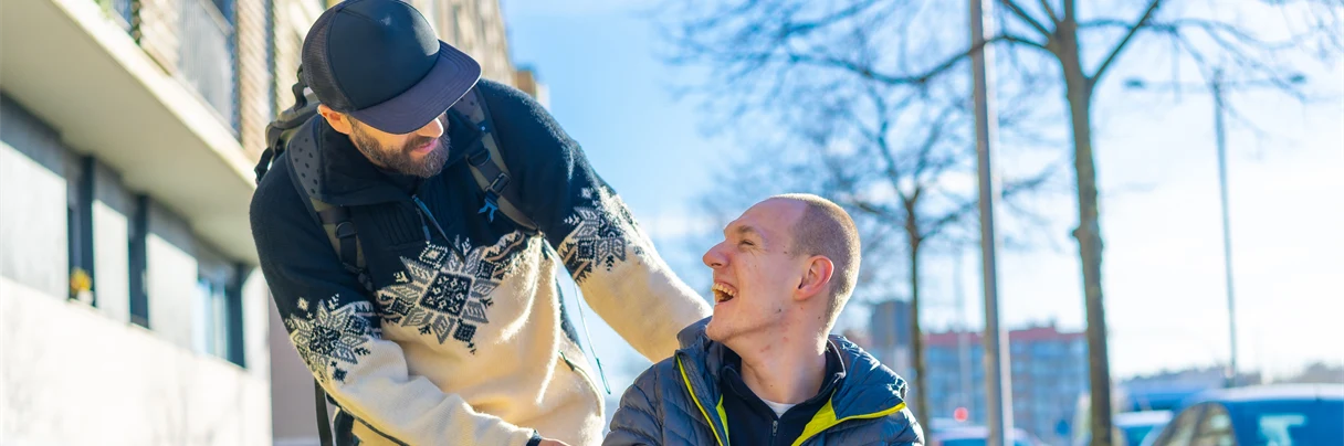 En man i rullstol och hans personliga assistans som tittar på varandra och ser glada ut.