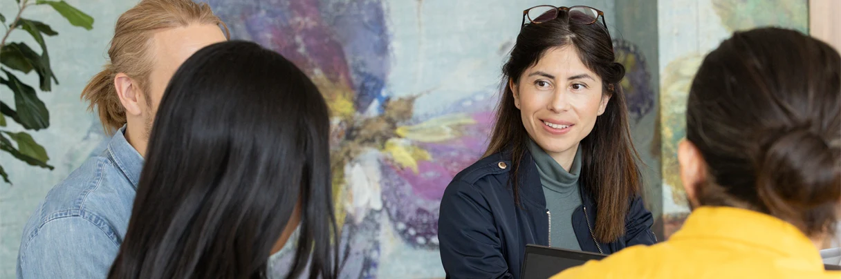 En grupp av människor sitter och pratar runt ett bord, en kvinna är vänd mot kameran, hon är glad 