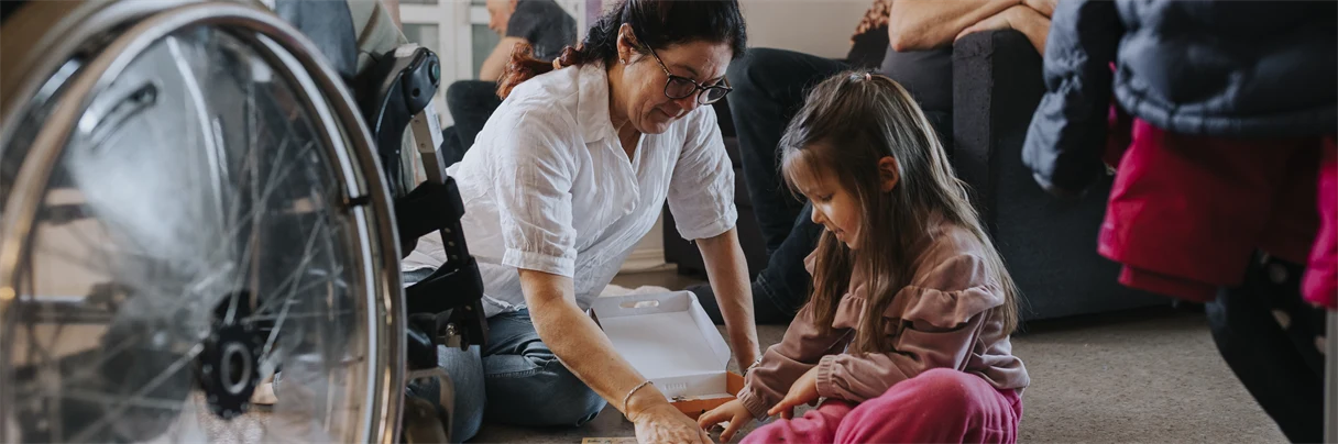 En flicka sitter på golvet tillsammans med en kvinna. En rullstol står parkerad bredvid dem. 