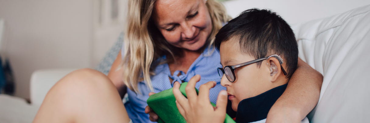 Leende mamma håller om pojke som använder läsplatta i soffa