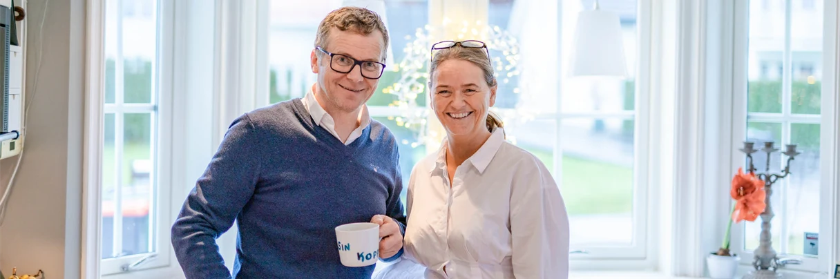 Mann og kone på kjøkkenet med kaffe kopp, han i blå genser og briller og hun smilende i hvit kjole