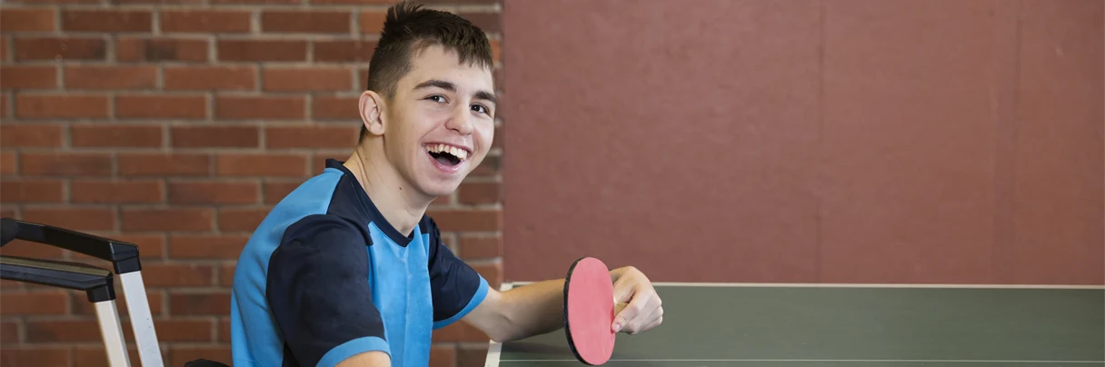 EN pojke sitter i en rullstol och spelar bordstennis. Pojken ler och tittar in i kameran.