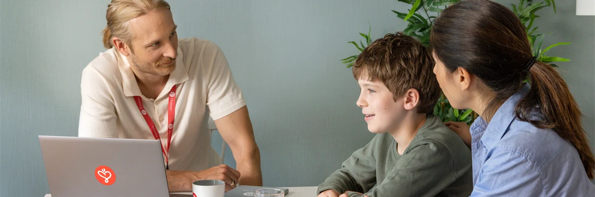 En man en kvinna och en pojke sitter vid ett bord, mannen har en dator och ett nyckelland med Humana logotypen på, de samtalar