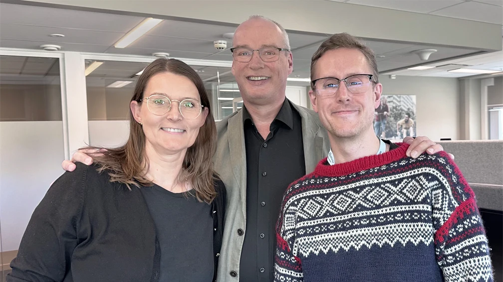 Tre smilende personer med briller på stående sammen på kontoret