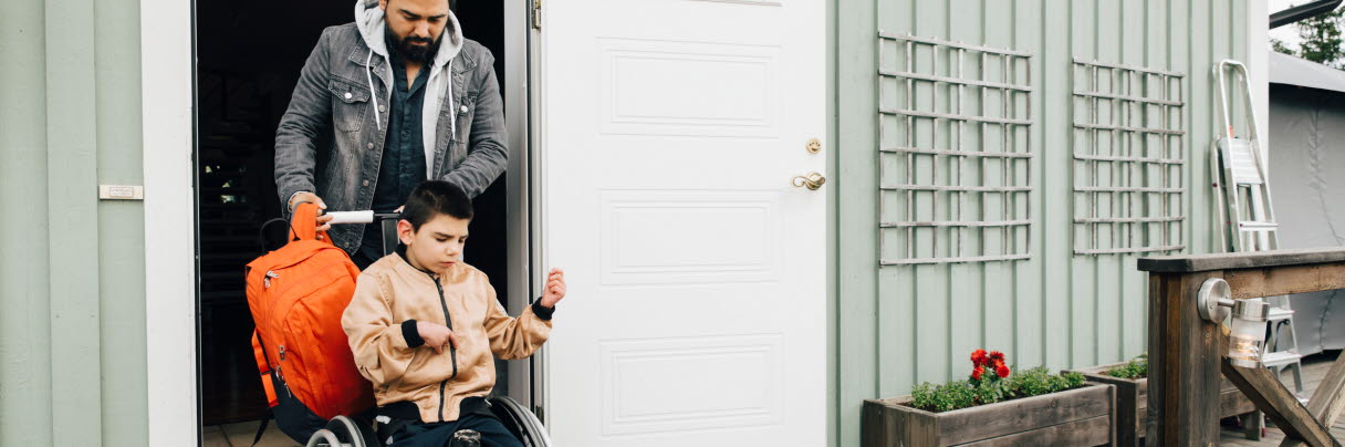En man skjutsar ut en rullstol med en pojke i från huset