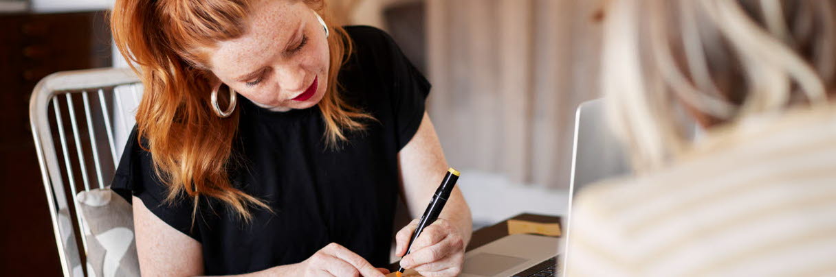 Kvinna med rött hår sitter vid ett bord och skriver med en penna. 