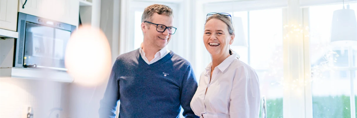 Mann og kone på kjøkkenet med kaffe kopp, han i blå genser og briller og hun smilende i hvit kjole