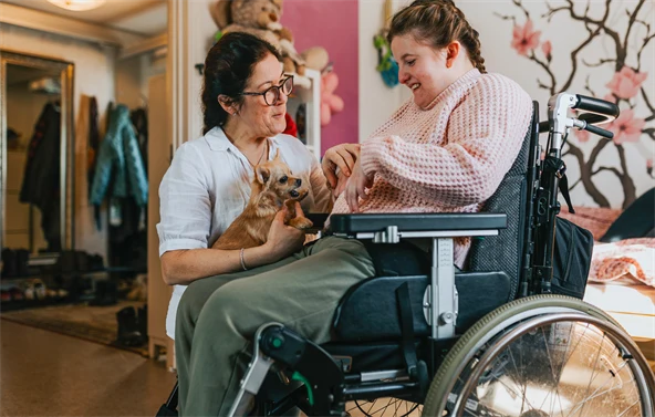 Kvinna i rullstol klappar en liten hund som assistenten håller i