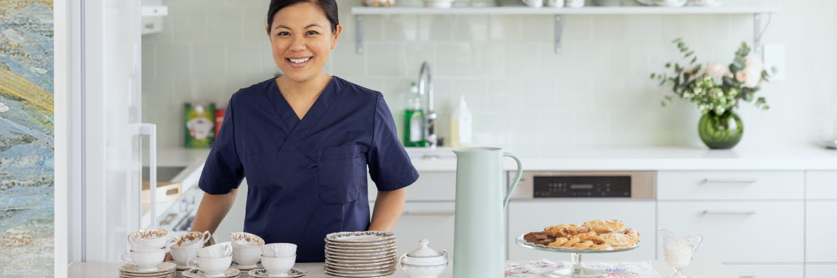 Kvinna med svart hår iklädd mörkblå tröja, står i ett kök med fika framför sig, hon är glad