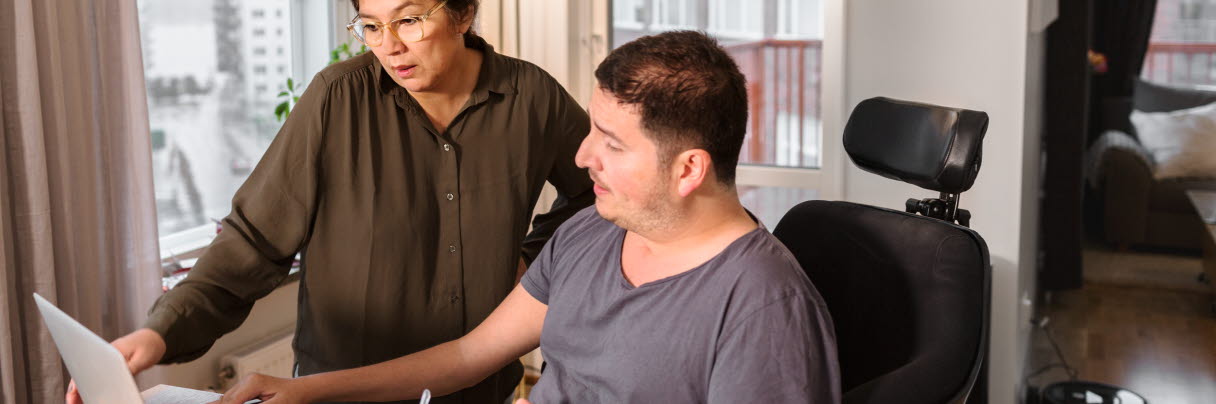 En man i rullstol tittar på dator tillsammans med sin assistent
