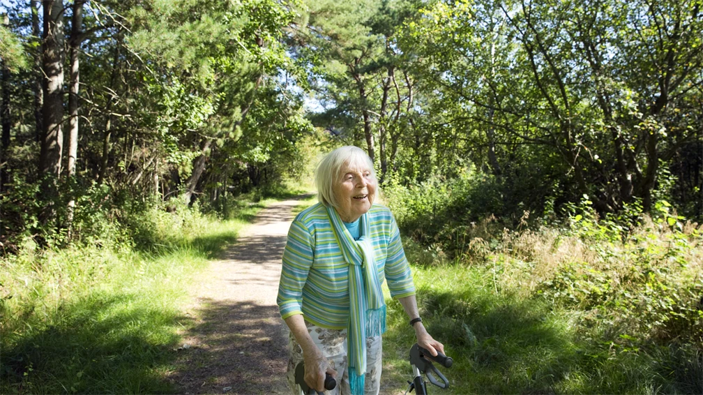En äldre kvinna med rullator går ute i naturen.