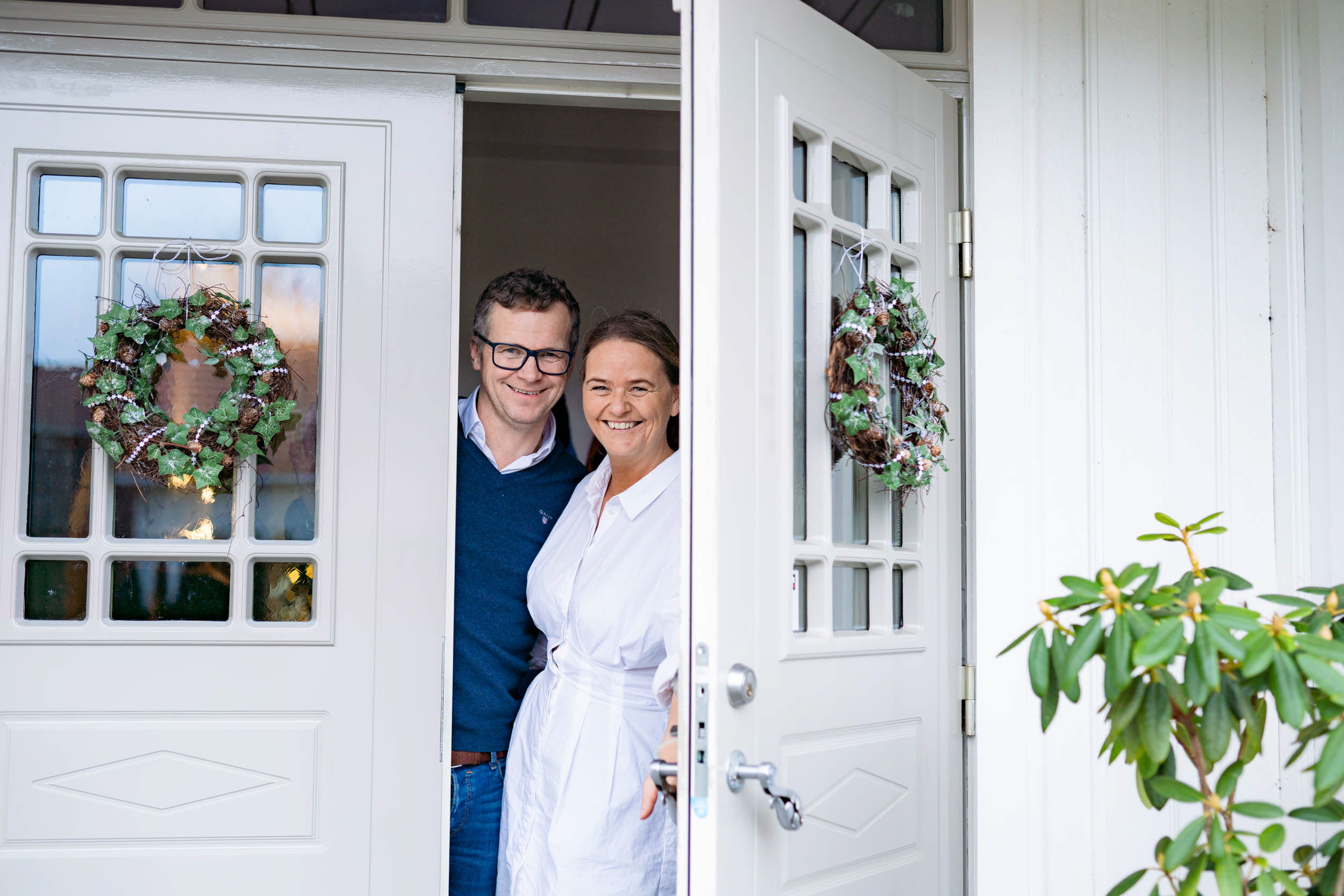 Mann og kone i inngangdøren, han i blå genser og briller og hun smilende i hvit kjole