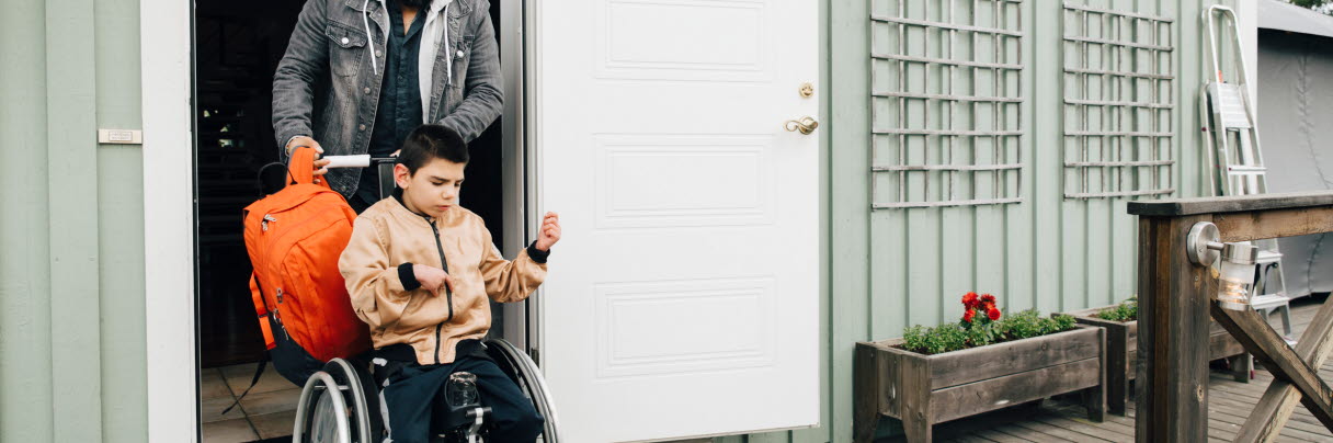En man skjutsar ut en rullstol med en pojke i från huset