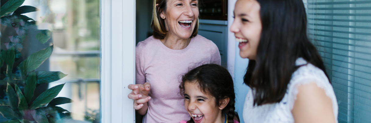 En äldre kvinna och två tjejer står i en balkongöppning och skrattar. 